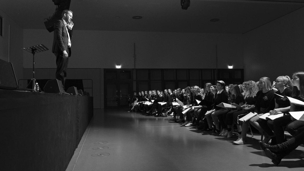 Der Dirigent steht auf der Bühne und spricht mit dem Jugendchor, der im Saal sitzt. Der Dirigent steht auf der Bühne und spricht mit dem Jugendchor, der im Saal sitzt.