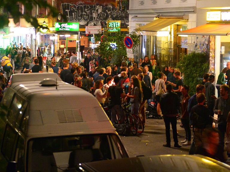 Junge Erwachsene stehen abends in einem Ausgehviertel auf der Straße und trinken.