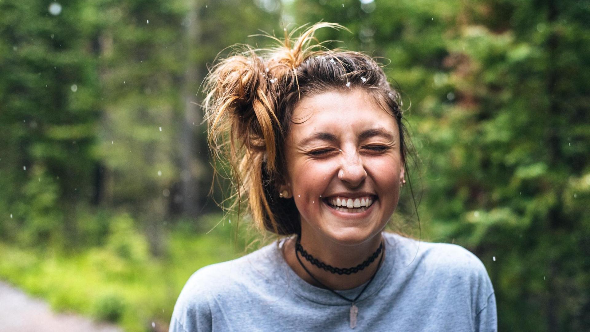 Schneeflocken fallen auf das Gesicht einer jungen Frau, die am Waldrand steht. Ihre Augen sind geschlossen und sie hat ein breites Lachen im Gesicht. Schneeflocken fallen auf das Gesicht einer jungen Frau, die am Waldrand steht. Ihre Augen sind geschlossen und sie hat ein breites Lachen im Gesicht.