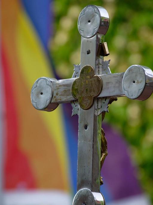 Ein Kreuz steht bei einem Open-Air-Segnungsgottesdienst für Liebende vor einer  Kirche. Im Hintergrund ist eine Regenbogenfahne zu sehen.