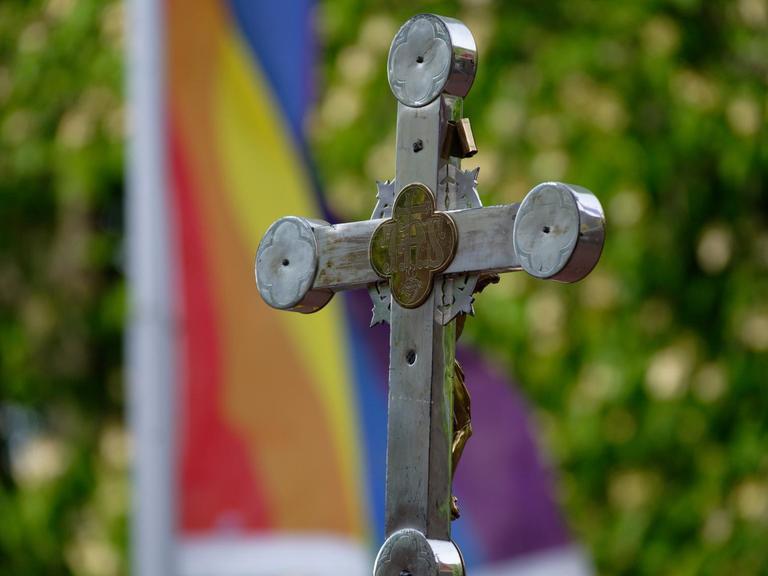 Ein Kreuz steht bei einem Open-Air-Segnungsgottesdienst für Liebende vor einer  Kirche. Im Hintergrund ist eine Regenbogenfahne zu sehen.