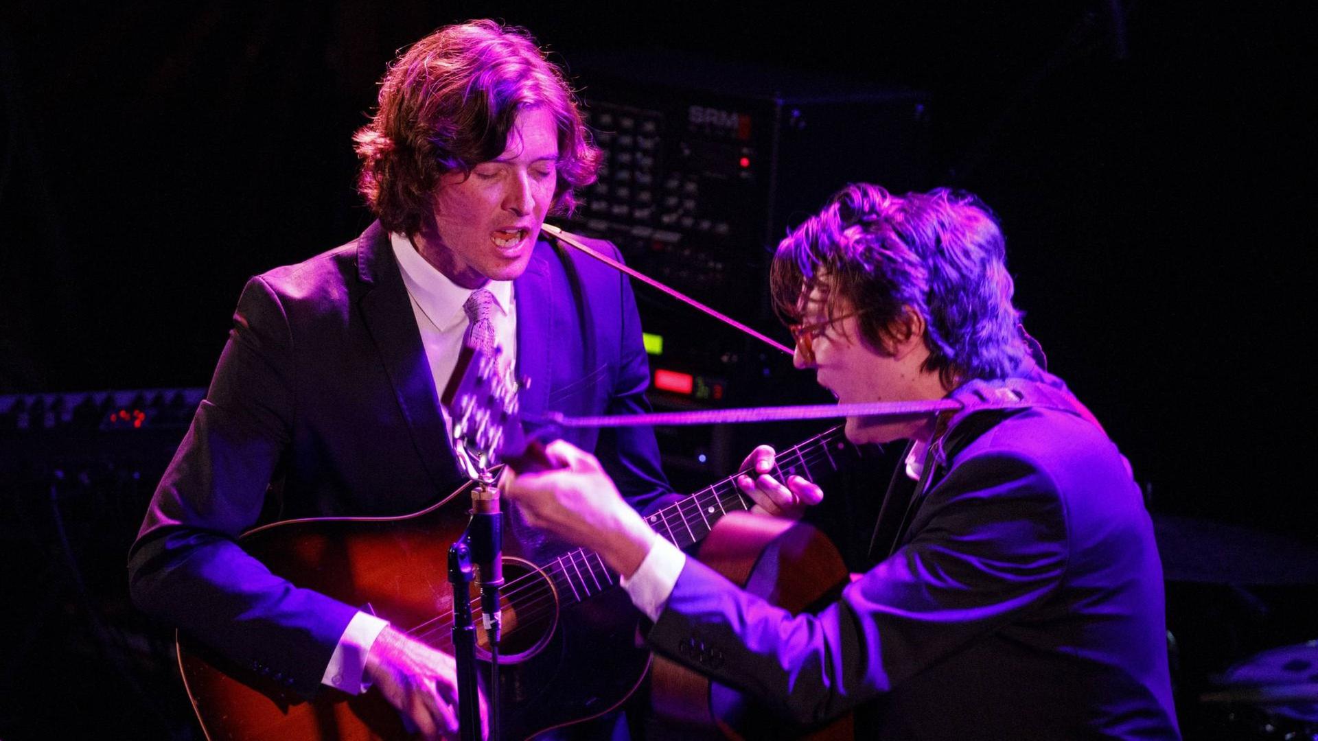Zwei Männer stehen sich mit Gitarren in lilafarbenem Licht gegenüber und singen.