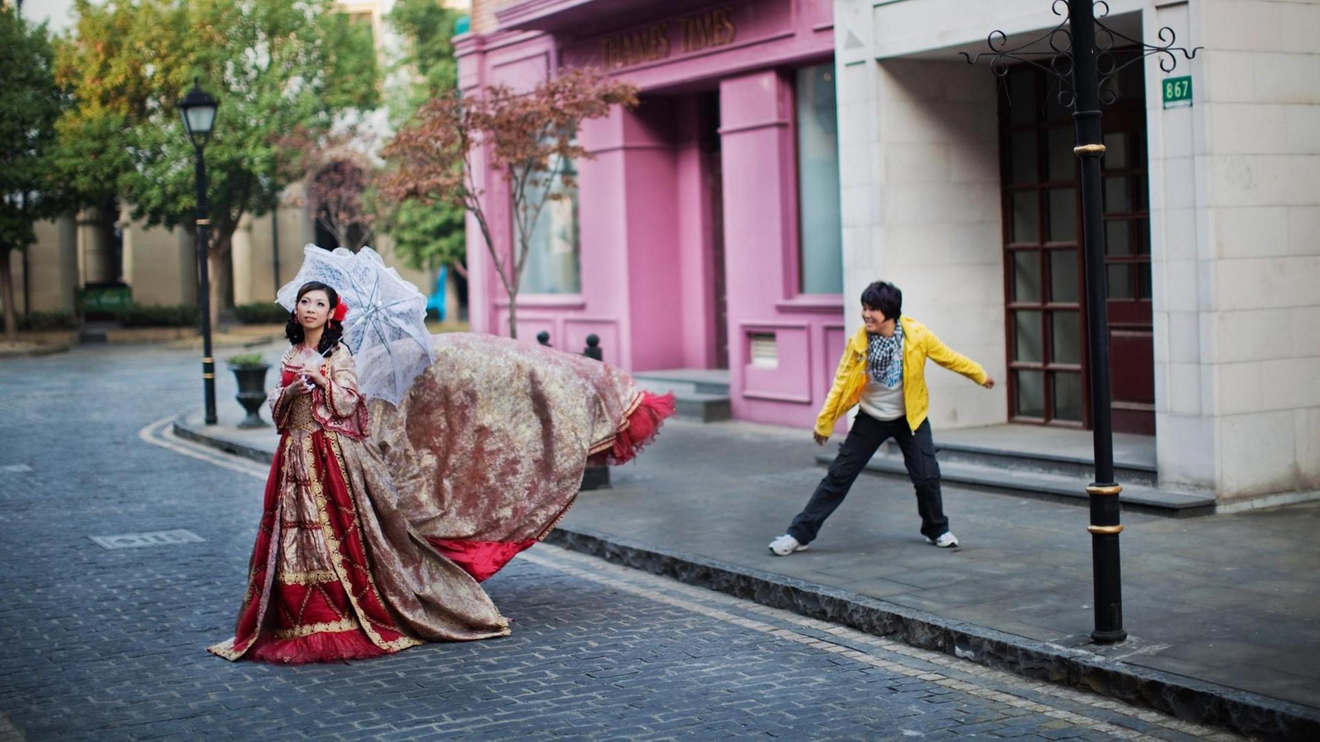 Ein chinesisches Paar posiert auf einer Strasse. Sie trägt einen Sonnenschirm und ein pompöses rotes Kleid. Er trägt eine knallgelbe Jacke. Ein chinesisches Paar posiert auf einer Strasse. Sie trägt einen Sonnenschirm und ein pompöses rotes Kleid. Er trägt eine knallgelbe Jacke.