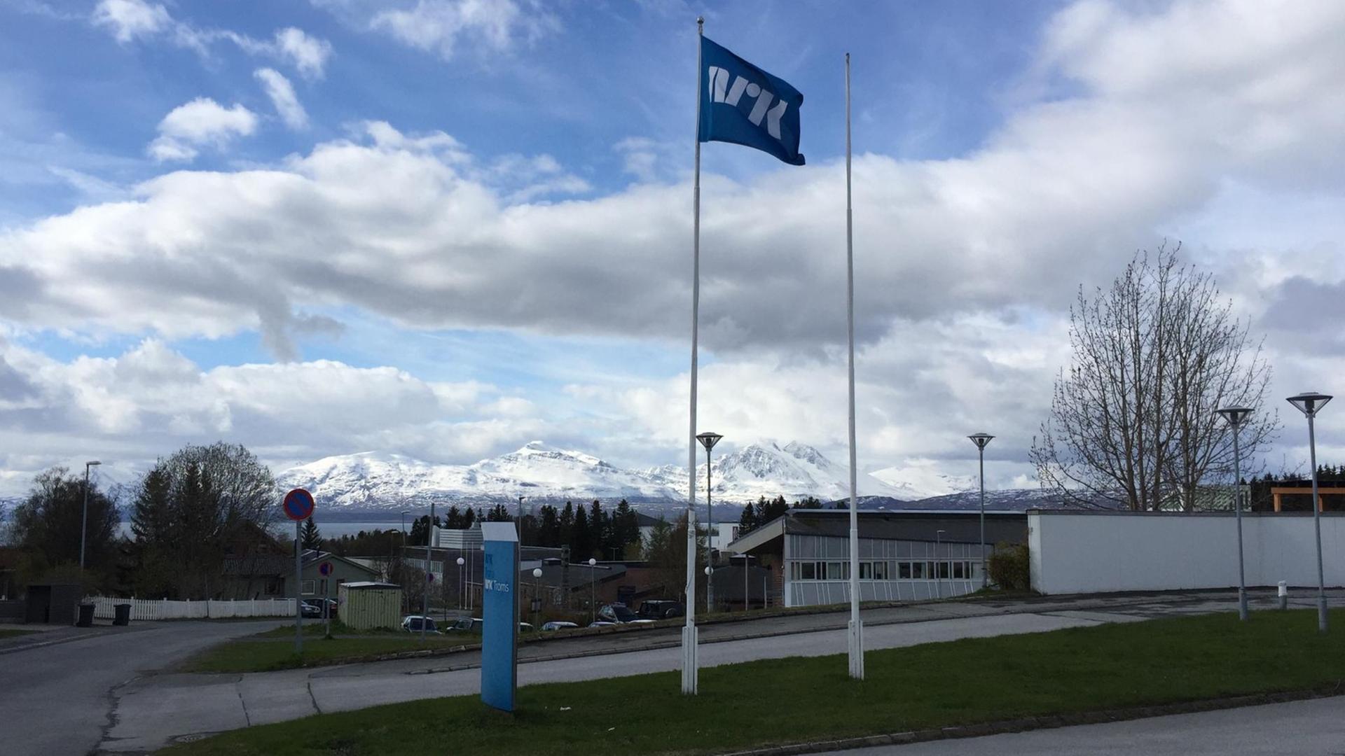Zu sehen ist eine Fahne vor dem Redaktionsgebäude des norwegischen Rundfunksenders NRK in Tromsø. Im Hintergrund sind schneebedeckte Berge zu erkennen. Zu sehen ist eine Fahne vor dem Redaktionsgebäude des norwegischen Rundfunksenders NRK in Tromsø. Im Hintergrund sind schneebedeckte Berge zu erkennen.