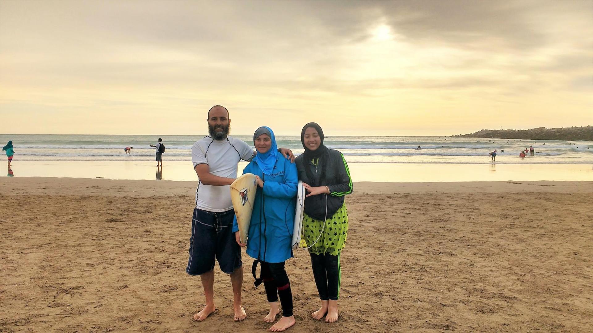 Aisha steht mit ihrem Vater und ihrer Cousine und den Surfboards am Strand