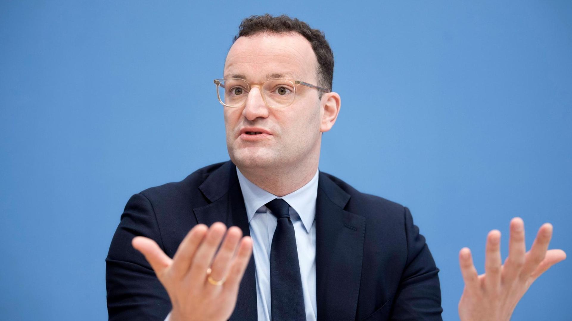 Bundesgesundheitsminister Jens Spahn bei einer Pressekonferenz: Der Minister steht in schwarzem Anzug, weißem Hemd und dunkler Krawatte vor einem mittelblauen Hintergrund und hält beide Handflächen offen nach vorn. Bundesgesundheitsminister Jens Spahn bei einer Pressekonferenz: Der Minister steht in schwarzem Anzug, weißem Hemd und dunkler Krawatte vor einem mittelblauen Hintergrund und hält beide Handflächen offen nach vorn.