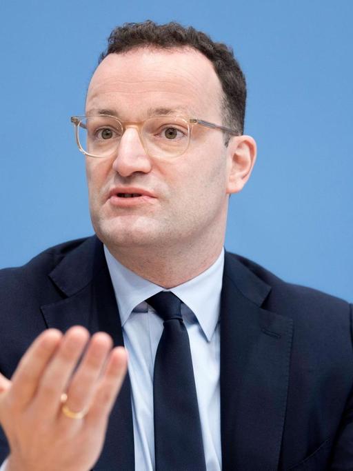 Bundesgesundheitsminister Jens Spahn bei einer Pressekonferenz: Der Minister steht in schwarzem Anzug, weißem Hemd und dunkler Krawatte vor einem mittelblauen Hintergrund und hält beide Handflächen offen nach vorn.