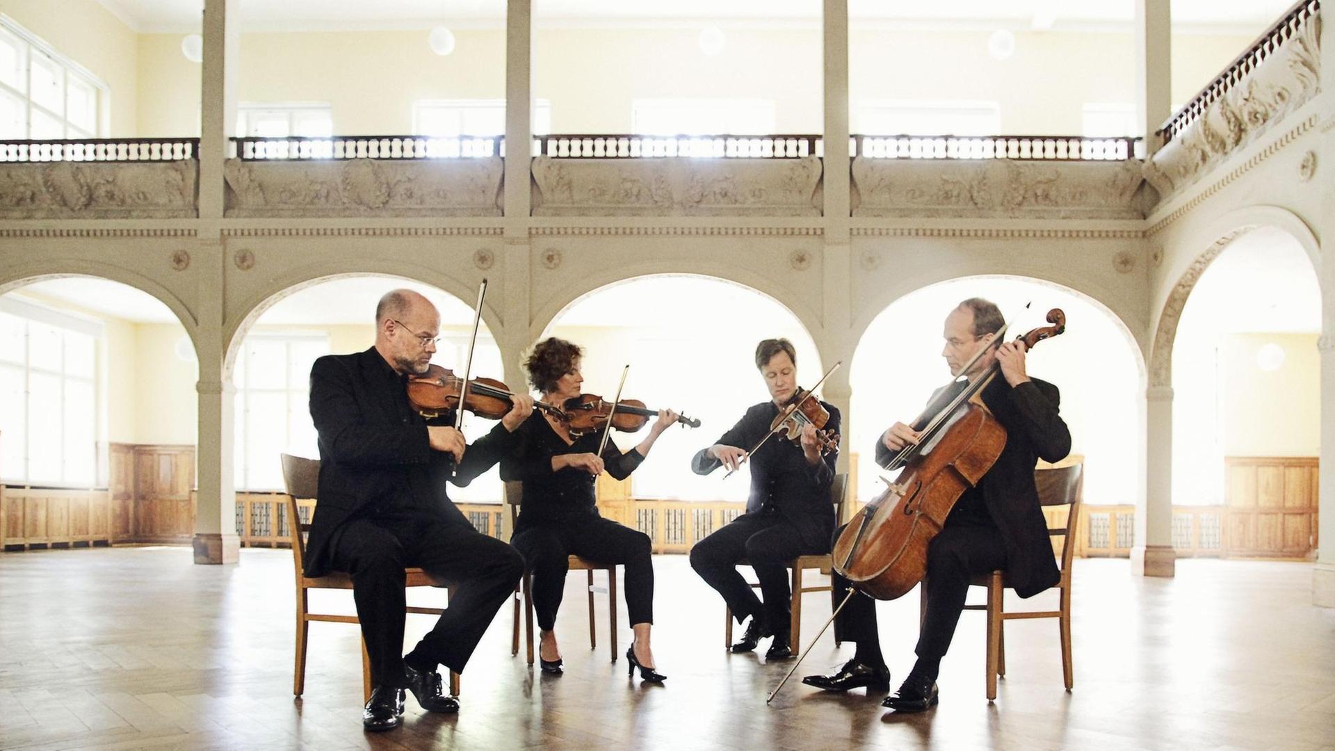 Eine Frau und drei Männer sitzen in einem hellen, hohen, lichtdurchfluteten historischen Raum und spielen gemeinsam Quartett. Eine Frau und drei Männer sitzen in einem hellen, hohen, lichtdurchfluteten historischen Raum und spielen gemeinsam Quartett.