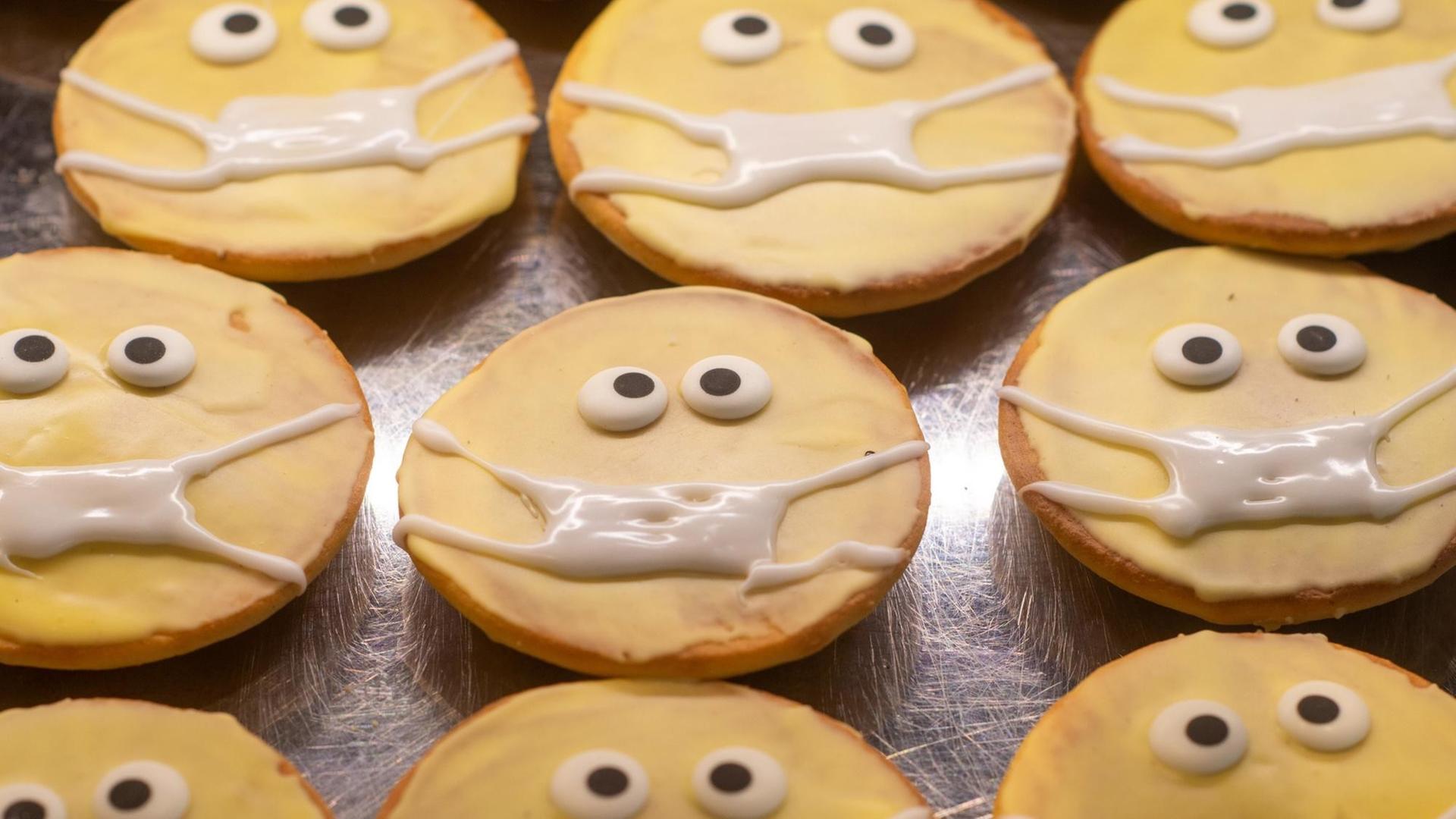 Kleine runde Kuchen in einer Auslage: Sie sind mit Zuckerguss verziert. Ihnen wurden zwei Augen und ein Mundschutz "aufgemalt". Kleine runde Kuchen in einer Auslage: Sie sind mit Zuckerguss verziert. Ihnen wurden zwei Augen und ein Mundschutz "aufgemalt".