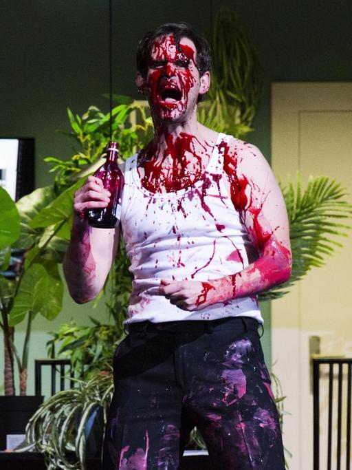 Ein Schauspieler im Unterhemd bespritzt sich mit einer blutroten Flüssigkeit aus einer Flasche.
