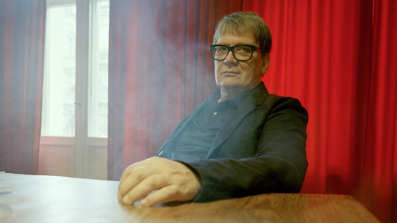 Sven Regener trägt eine schwarze Brille und einen schwarzen Anzug und sitzt an einem Holztisch vor einem Fenster mit roten Vorhängen. Es steigt Rauch von unter dem Tisch auf. Sven Regener trägt eine schwarze Brille und einen schwarzen Anzug und sitzt an einem Holztisch vor einem Fenster mit roten Vorhängen. Es steigt Rauch von unter dem Tisch auf.