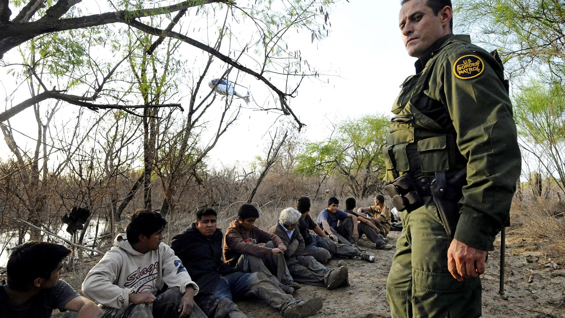Ein Grenzschützer in Uniform steht vor einer Reihe Menschen, die am Boden sitzen. Im Hintergrund ist Wasser zu sehen.