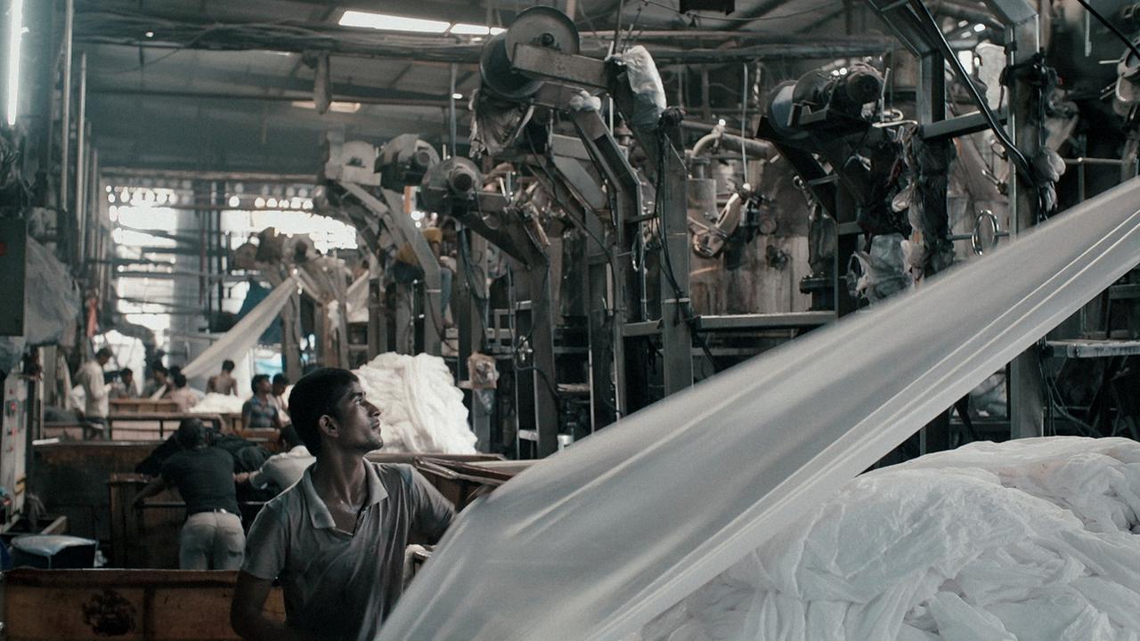 Szenenbild aus "Machines": Männer arbeiten an großen Maschinen in der düsteren Fabrikhalle