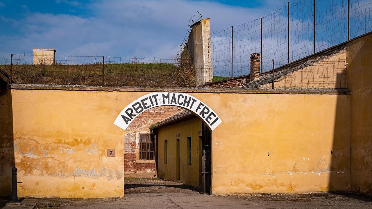 Eine gelbe Mauer einer ehemaligen Festung mit einem halbrunden Torbogen, der den Schriftzug "Arbeit macht frei" trägt und der Eingang zum Konzentrationslager Theresienstadt war.