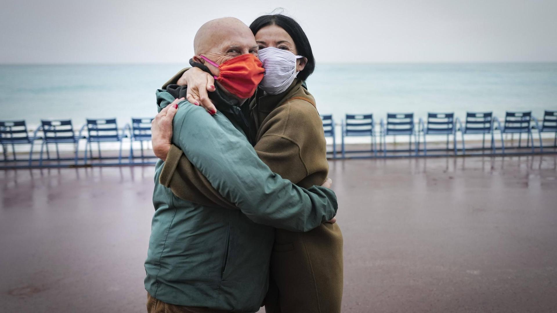 Ein älteres Pärchen umarmt sich auf einer Strandpromenade mit Masken und schaut in die Kamera Ein älteres Pärchen umarmt sich auf einer Strandpromenade mit Masken und schaut in die Kamera