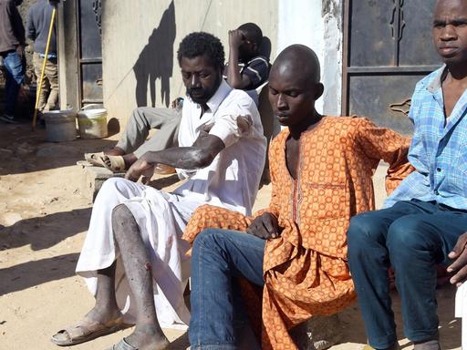 Männer aus afrikanischen Staaten sitzen an einer Häuserwand auf einer unbefestigten Straße.