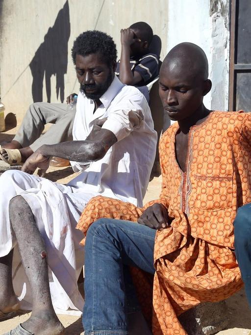 Männer aus afrikanischen Staaten sitzen an einer Häuserwand auf einer unbefestigten Straße.