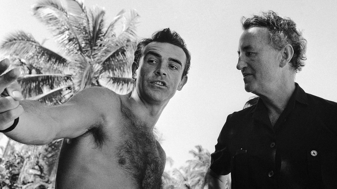 Schwarzweißfoto von Sean Connery mit nacktem Oberkörper und dem Schriftsteller Ian Fleming unter freiem Himmel. Schwarzweißfoto von Sean Connery mit nacktem Oberkörper und dem Schriftsteller Ian Fleming unter freiem Himmel.