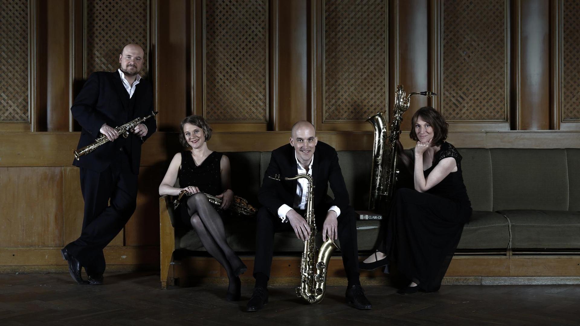 Vier Musiker sitzen mit ihren unterschiedlich großen Saxophonen in Abendrobe auf einer Polsterbank in einem mit dunklem, schweren Holz vertäfelten Raum und schauen lachend in die Kamera. Vier Musiker sitzen mit ihren unterschiedlich großen Saxophonen in Abendrobe auf einer Polsterbank in einem mit dunklem, schweren Holz vertäfelten Raum und schauen lachend in die Kamera.