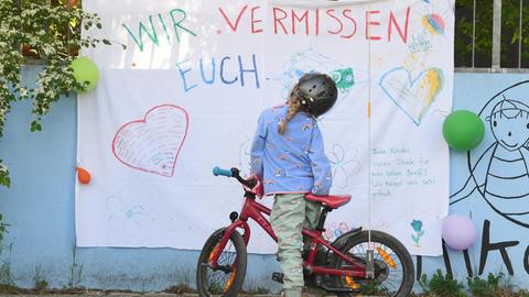 Ein Mädchen auf dem Fahrrad steht vor der Fassade einer Kita mit der Aufschrift "Wir vermissen euch".
