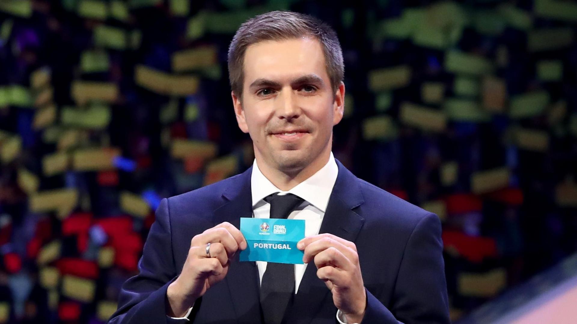 Lahm im dunkelblauen Anzug hält lächelnd das Los mit der Aufschrift "Final Draw - Portugal" in die Kamera. Lahm im dunkelblauen Anzug hält lächelnd das Los mit der Aufschrift "Final Draw - Portugal" in die Kamera.