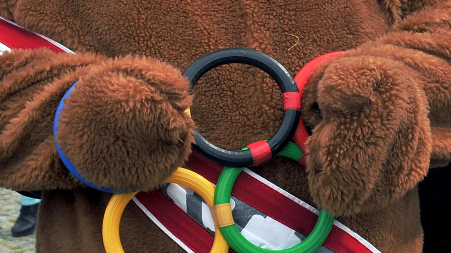 Ein als Bär verkleideter Mann streckt die Hände durch symbolische Olympische Ringe. Ein als Bär verkleideter Mann streckt die Hände durch symbolische Olympische Ringe.