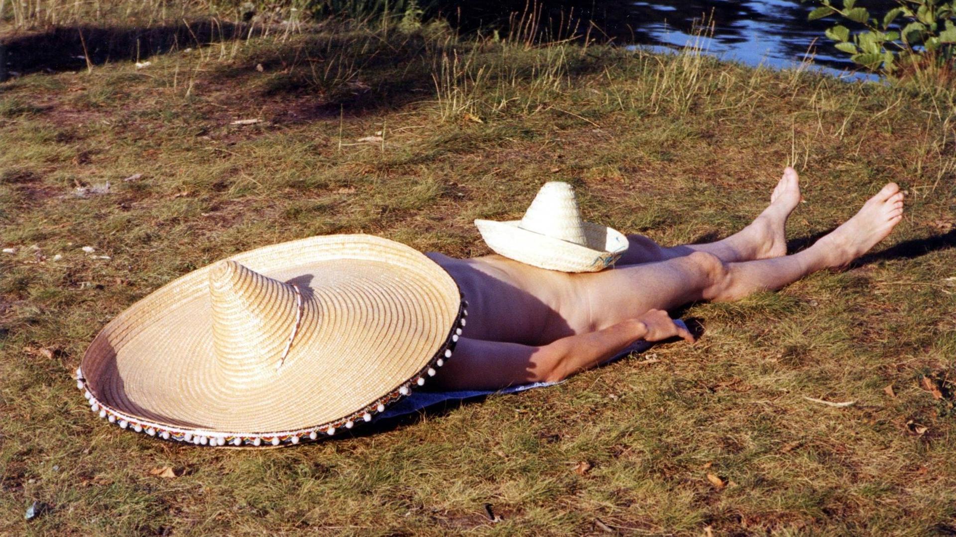 Ein Mann sonnt sich nackt. Über seinem Kopf und seinem Intimbereich liegt jeweils ein Sombrero.