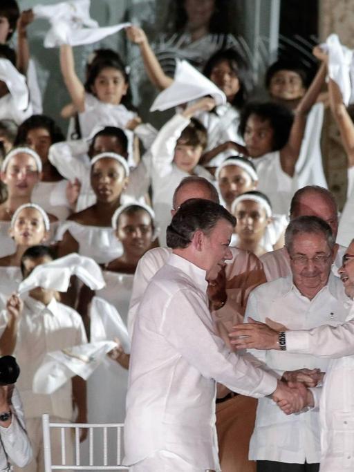Drei Männer in weißen Händen schütteln sich die Hände, umgeben von vielen anderen Männern und Frauen, die Fahnen schwingen und auch in Weiß gekleidet sind.