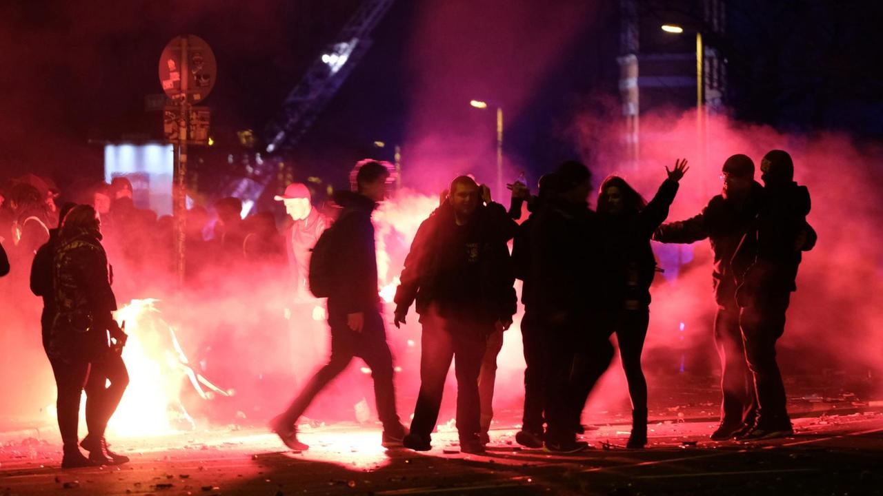 Eine Gruppe von Feiernden in der Silvesternacht steht im Schein von Böllern und Raketen auf der Straße. Eine Gruppe von Feiernden in der Silvesternacht steht im Schein von Böllern und Raketen auf der Straße.