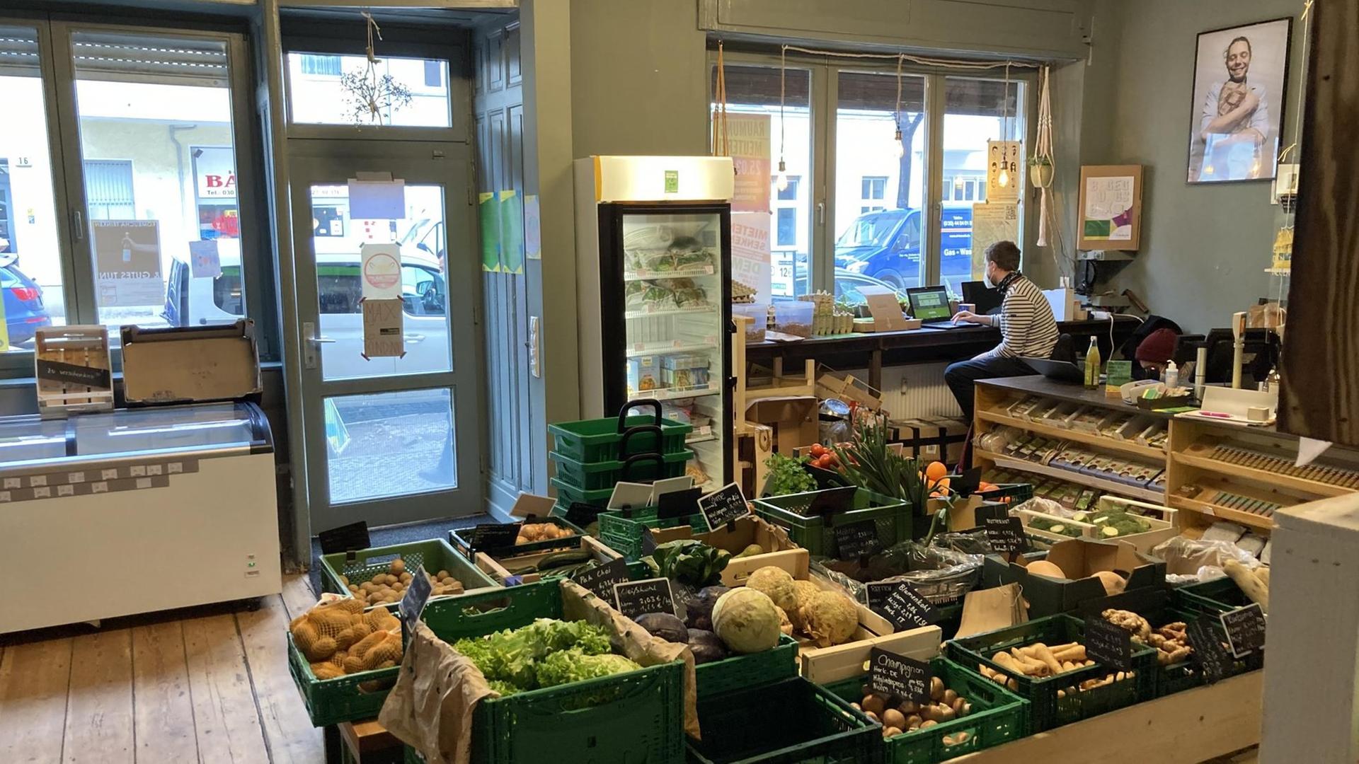 In einem kleinen Bioladen mit Holzfußboden ist Gemüse in grünen Kisten aufgestellt. In der Ecke sitzt ein junger Mann an einer Kasse. In einem kleinen Bioladen mit Holzfußboden ist Gemüse in grünen Kisten aufgestellt. In der Ecke sitzt ein junger Mann an einer Kasse.