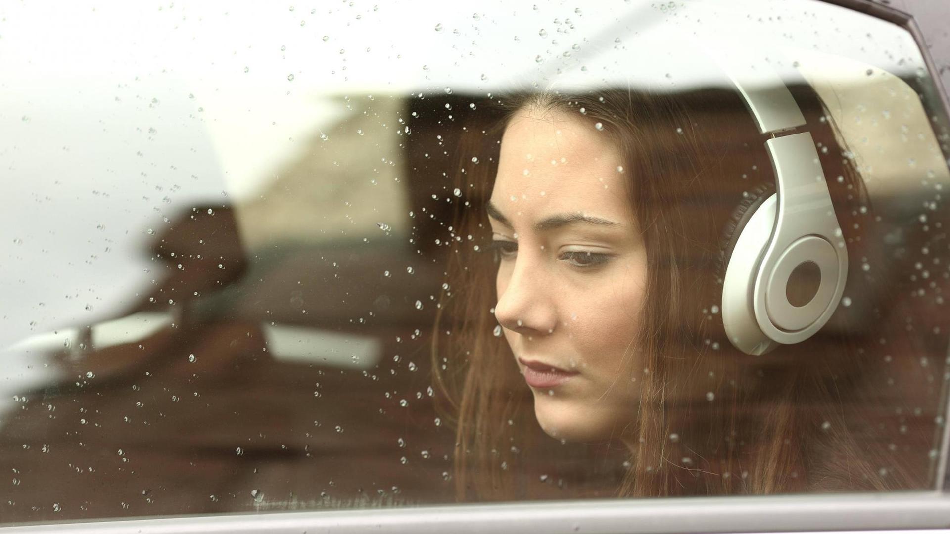 Eine junge Frau mit Kopfhörern blickt gedankenverloren aus einem Autofenster. Eine junge Frau mit Kopfhörern blickt gedankenverloren aus einem Autofenster.