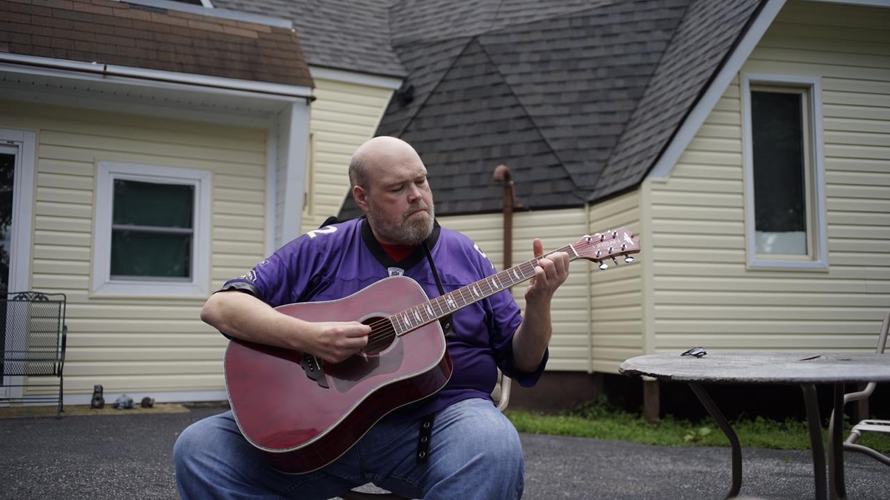 Mann mit lila Shirt, spielt Gitarre.