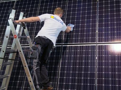 Ein Mitarbeiter einer Dresdner Solarfirma steht auf einer Leiter und legt letzte Hand an einer Photovoltaikanlage an, bei der die Solarzellen senkrecht an einer Wand angebracht sind.