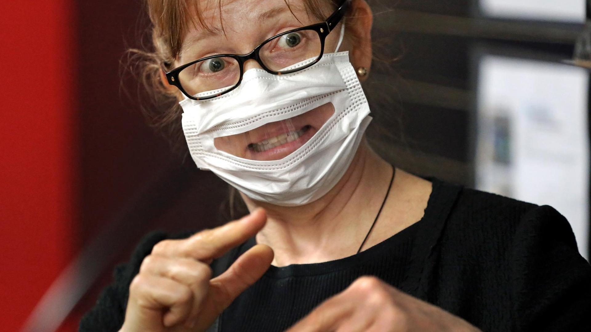 Eine Frau trägt eine Maske mit einem Sichtfenster und macht eine Geste aus der Gebärdensprache Eine Frau trägt eine Maske mit einem Sichtfenster und macht eine Geste aus der Gebärdensprache