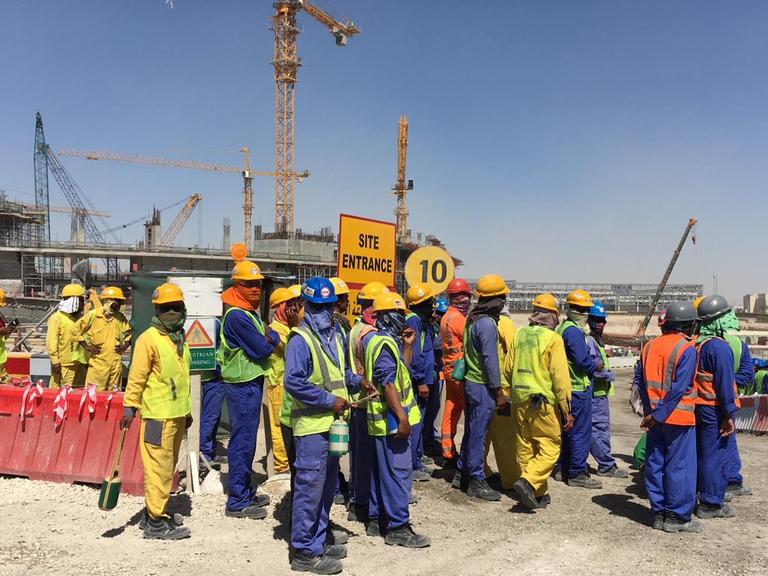 Bauarbeiter stehen auf riesiger Baustelle, das Gesicht mit Tüchern verpackt gegen die Hitze