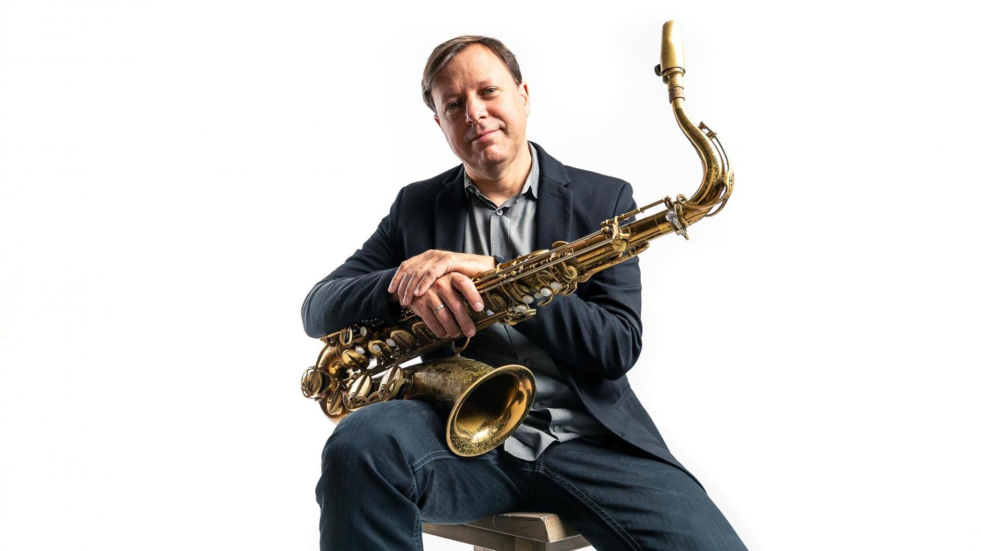 Ein Mann sitzt vor weißem Hintergrund auf einen Hocker und hält schmunzelnd ein Saxofon in beiden Händen