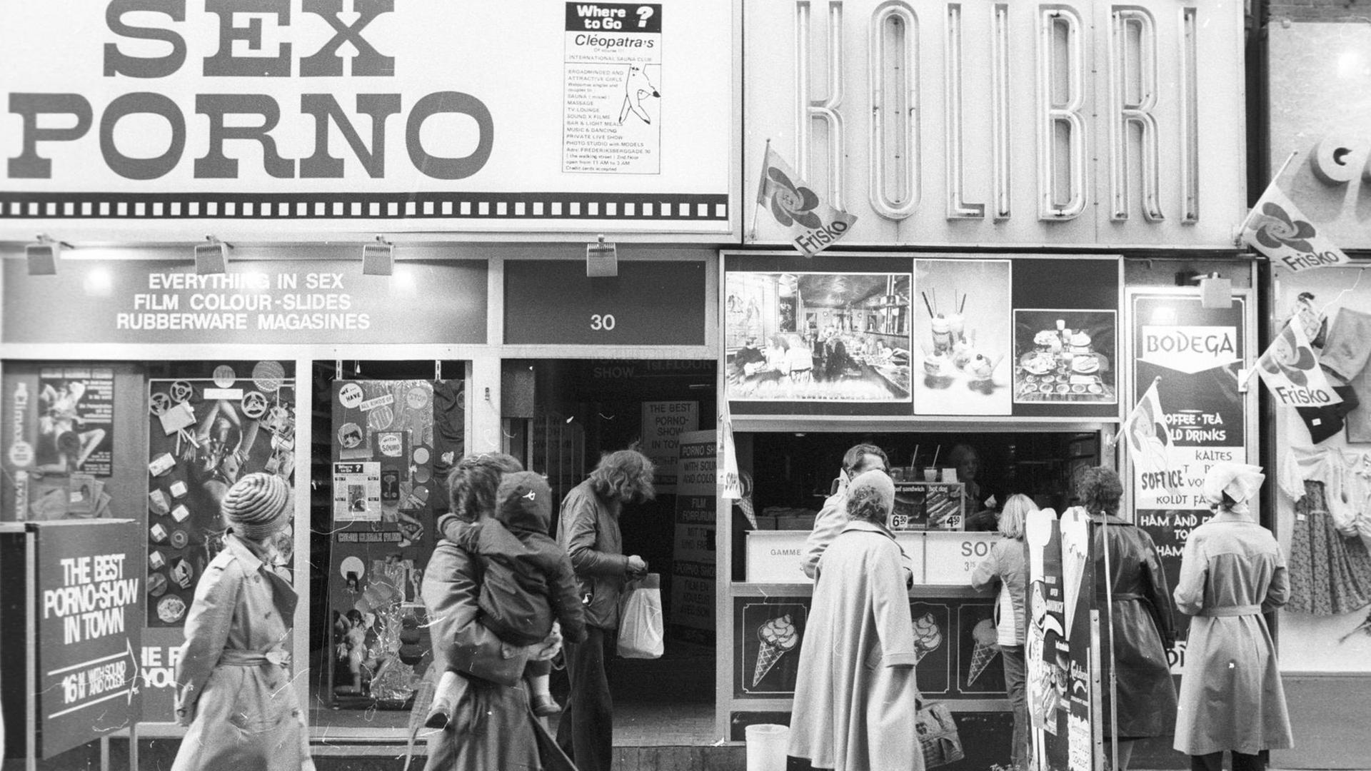 Das Bild zeigt eine Straße in Kopenhagen mit vorbeilaufenden Menschen. Auf der linken Seite ist ein Laden mit der großen Aufschrift: "Sex Porno" zu sehen. Das Bild zeigt eine Straße in Kopenhagen mit vorbeilaufenden Menschen. Auf der linken Seite ist ein Laden mit der großen Aufschrift: "Sex Porno" zu sehen.