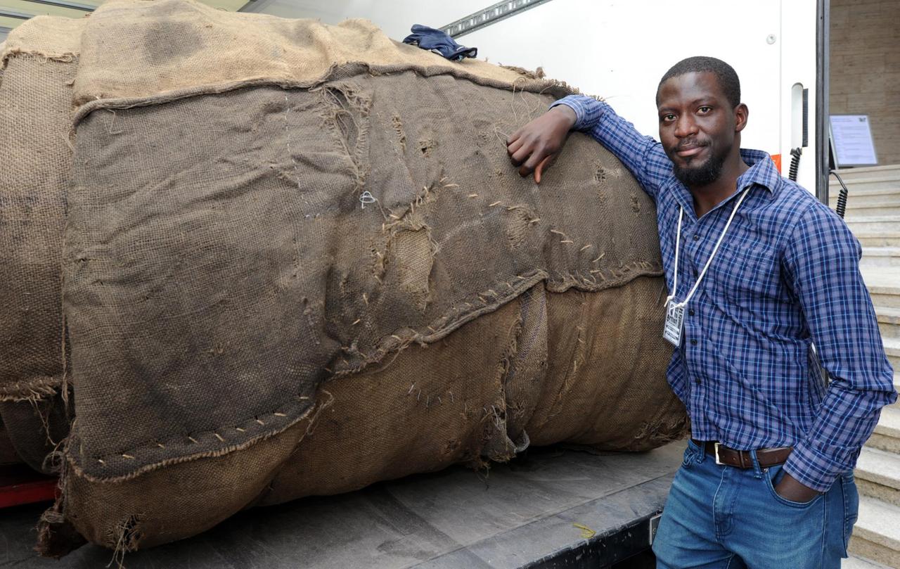Der documenta-Künstler Ibrahim Mahama aus Ghana lächelt am 23.05.2017 während der Aufbauarbeiten für die Verhüllung des Torhauses in Kassel (Hessen) neben zusammengenähten Jutesäcken zur documenta 14 in Kassel. Der documenta-Künstler Ibrahim Mahama aus Ghana lächelt am 23.05.2017 während der Aufbauarbeiten für die Verhüllung des Torhauses in Kassel (Hessen) neben zusammengenähten Jutesäcken zur documenta 14 in Kassel.