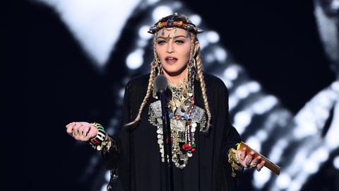 Das Bild zeigt die Sängerin Madonna während einer Rede. Sie trägt ein schwarzes Gewand mit vielen Lagen silbernen Schmuck um ihren Hals und um ihre Handgelenke. Auf dem Kopf trägt sie eine Art Krone mit spitzen Zacken.
