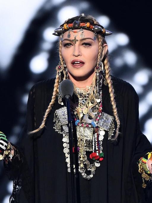 Das Bild zeigt die Sängerin Madonna während einer Rede. Sie trägt ein schwarzes Gewand mit vielen Lagen silbernen Schmuck um ihren Hals und um ihre Handgelenke. Auf dem Kopf trägt sie eine Art Krone mit spitzen Zacken.