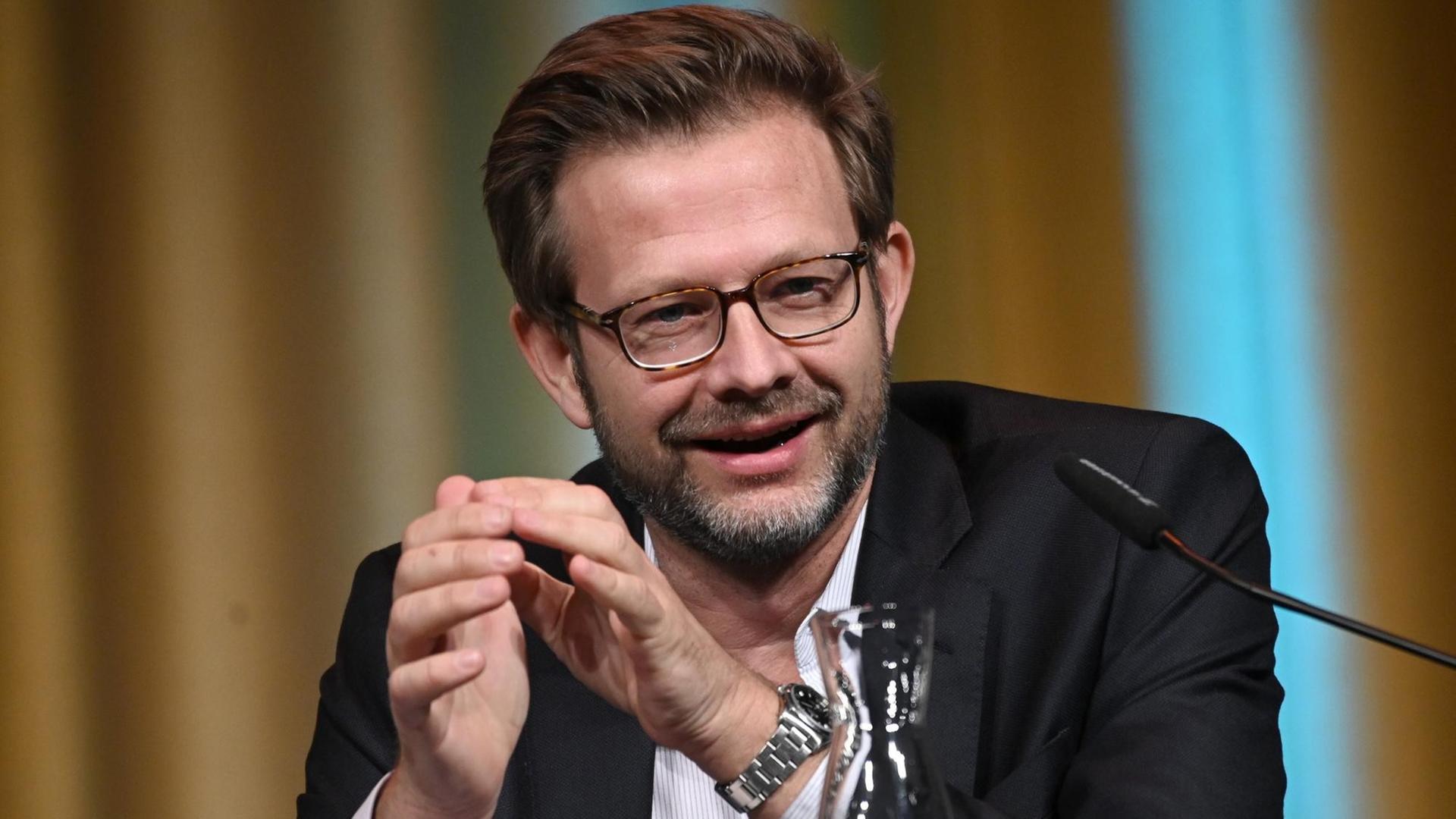 Der Autor Florian Illies sitzt bei Lesung an einem Tisch, auf dem ein Glas und eine Flasche stehen. Der Autor Florian Illies sitzt bei Lesung an einem Tisch, auf dem ein Glas und eine Flasche stehen.