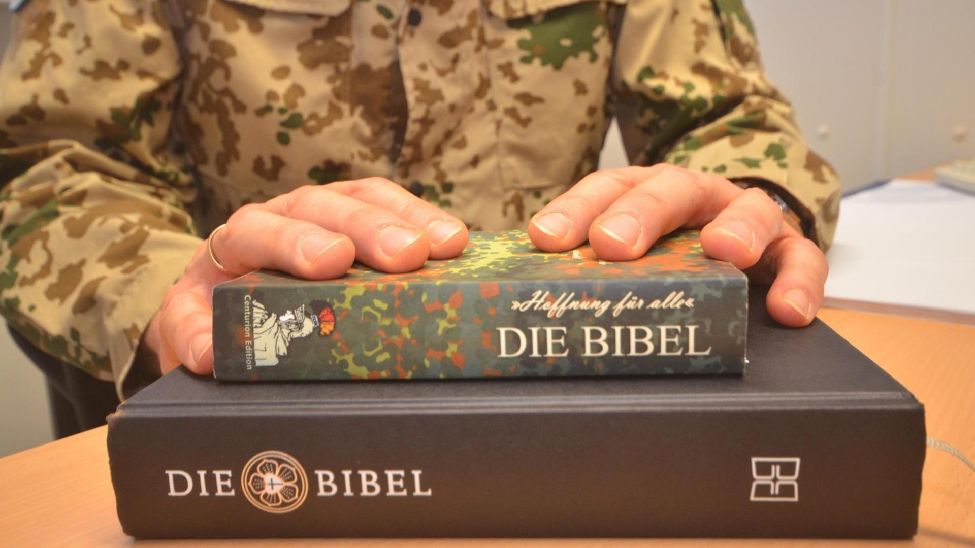 Eine Bibel in Flecktarn liegt auf einer klassisch gebundenen Bibel - die Hände des Militärpfarrers darüber. Eine Bibel in Flecktarn liegt auf einer klassisch gebundenen Bibel - die Hände des Militärpfarrers darüber.