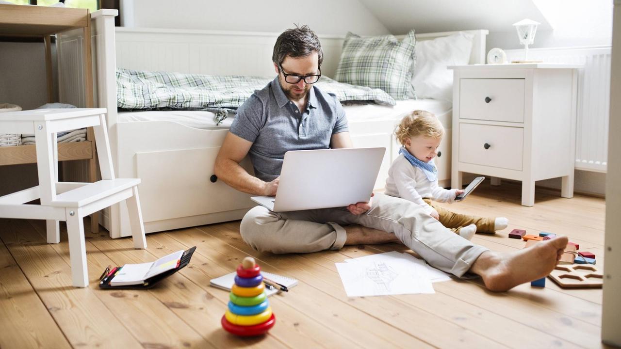 Ein Vater arbeitet am Laptop, während sein Kind daneben spielt.