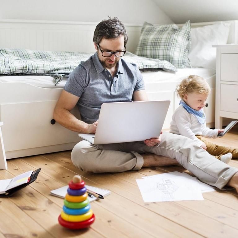 Ein Vater arbeitet am Laptop, während sein Kind daneben spielt.