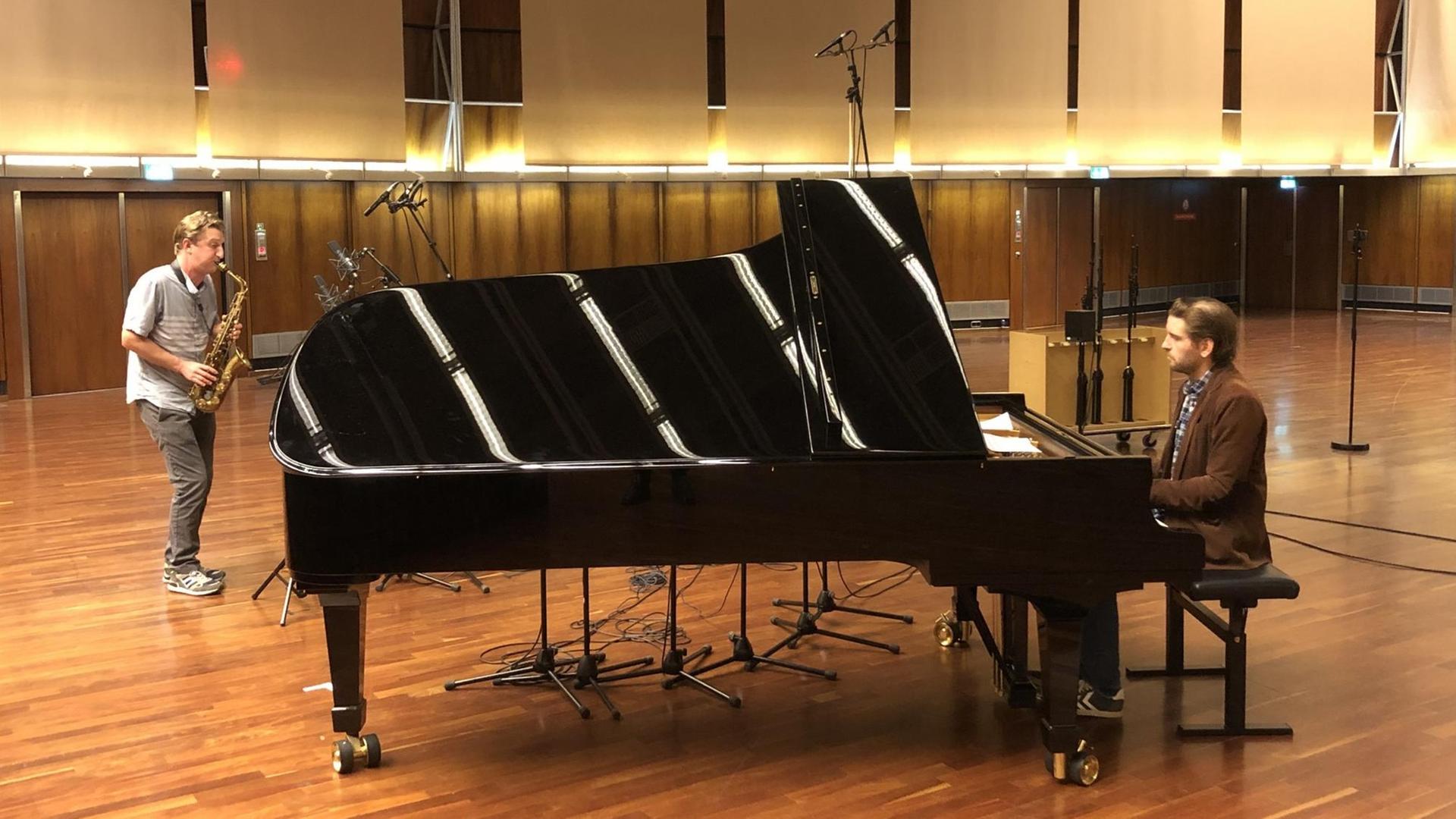 Zwei Musiker in Alltagskleidung spielen in einem Saal mit Parkettboden. Der rechte sitzt am schwarzen, aufgeklappten Konzertflügel, der den größten Teil des Bildes einnimmt. Links davon ein Altsaxofonist. Zwei Musiker in Alltagskleidung spielen in einem Saal mit Parkettboden. Der rechte sitzt am schwarzen, aufgeklappten Konzertflügel, der den größten Teil des Bildes einnimmt. Links davon ein Altsaxofonist.