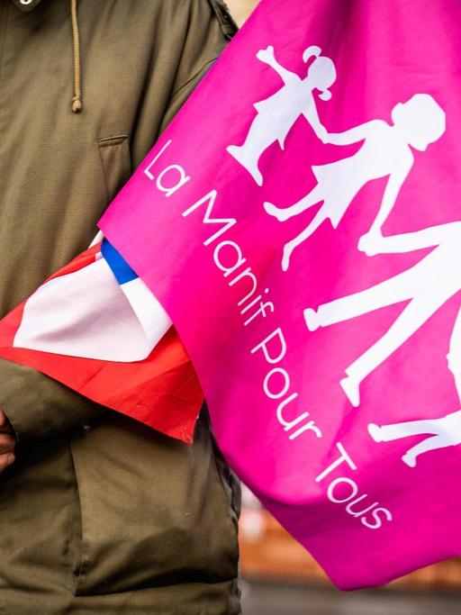 Detail der gekreuzten Hände eines Demonstranten mit einer französischen Flagge und einer Flagge der Demonstration mit einer Darstellung einer Normalfamilie mit Vater, Mutter und zwei Kindern.