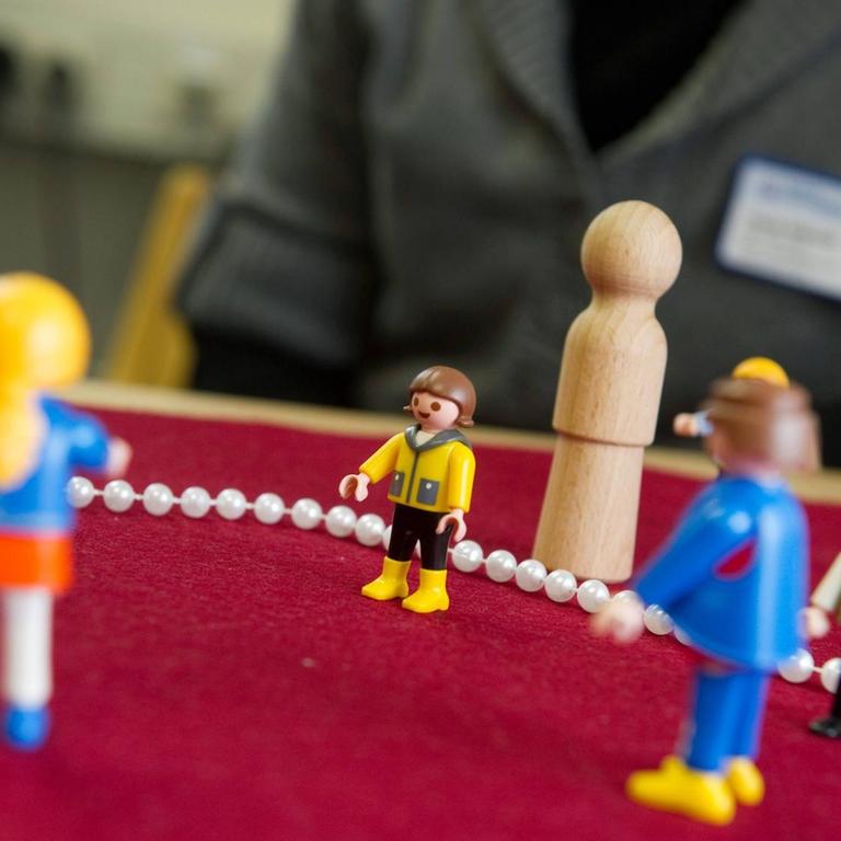 Zu sehen sind mehrere Playmobil-Figuren sowie einfache Holz-Figuren, die von einer Person auf einem Tisch aufgestellt werden.