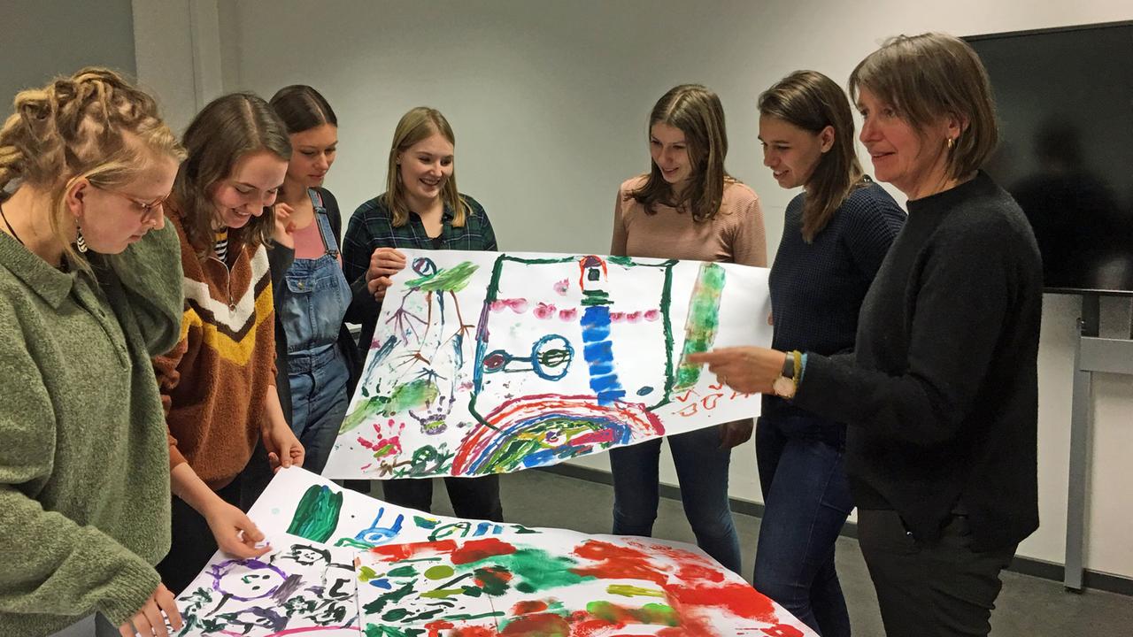 Professorin Karin Borck und ihre Studentinnen halten Bilder mit Trostwesen hoch, die Kinder gemalt haben.