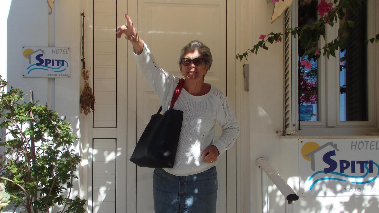Gisela steht vor einem griechischen Haus und schaut fröhlich in die Kamera