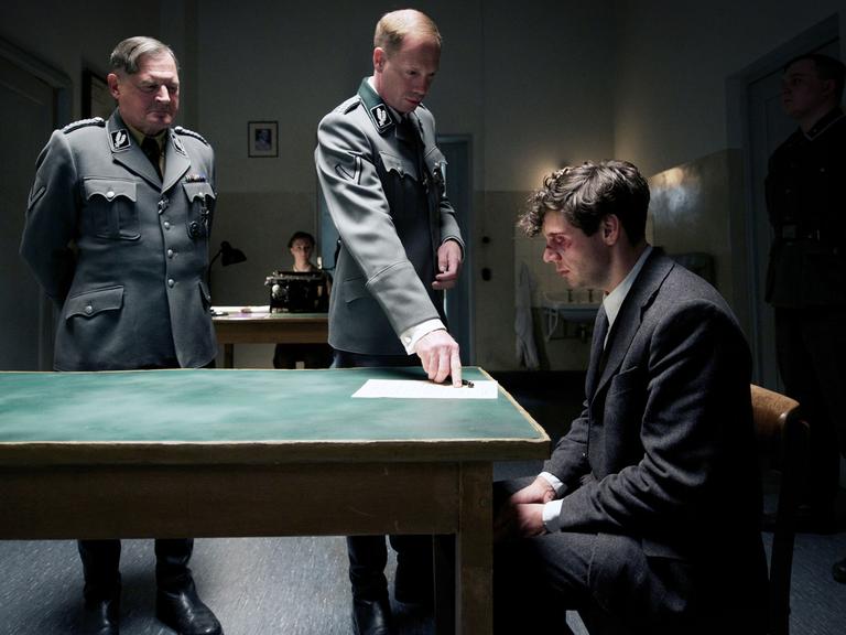 Burghart Klaußner (l) als Arthur Nebe und Johann von Bülow (M) als Heinrich Müller drängen Christian Friedel als Georg Elser in einer Szene des Kinofilms "Elser" ein Verhörprotokoll zu unterzeichnen.
